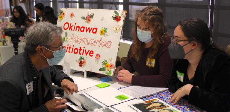 Two woman talking to a man at a booth that say Okinawa Memories Initiative.
