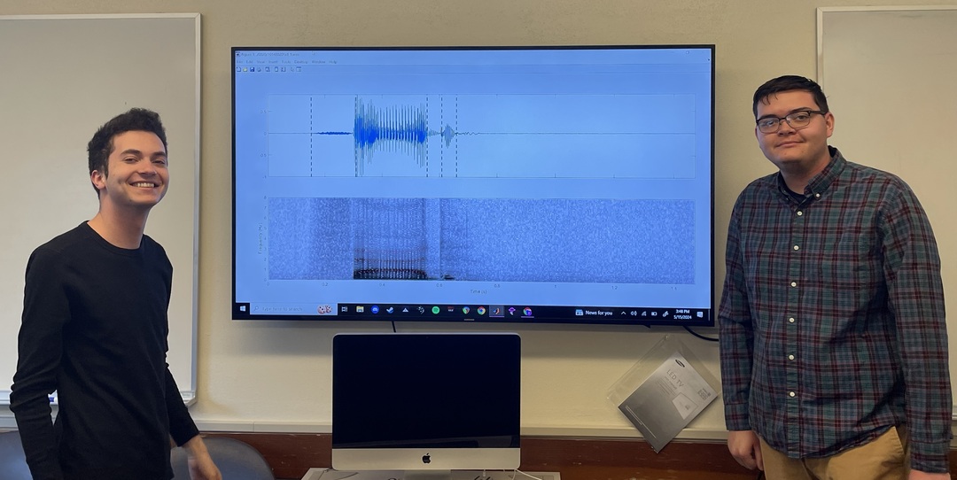 Two students standing in front on a tv with an audio file displayed